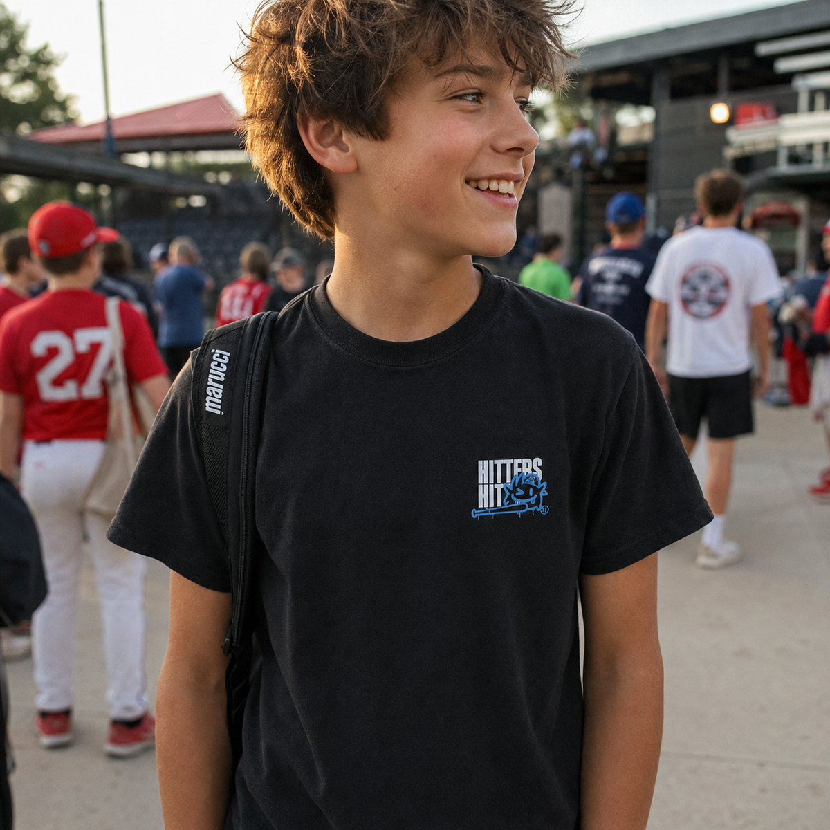 Boy wearing black youth baseball T-shirt with small “Hitters Hit” chest graphic at the ballpark.