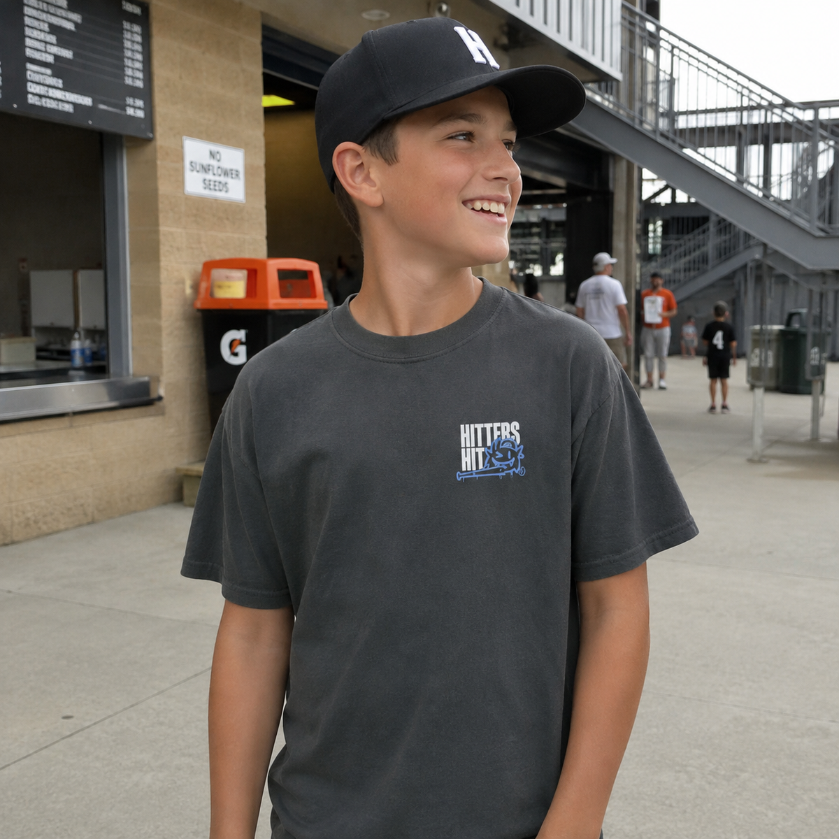 Boy wearing pepper youth baseball T-shirt with small “Hitters Hit” chest graphic at a baseball field.