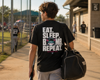Teen walking through a baseball complex in a black adult baseball T-shirt with the large “Eat. Sleep. Bomb. Repeat.” back graphic.