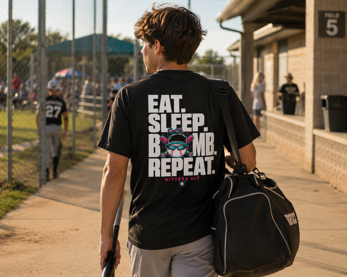 Teen walking through a baseball complex in a black adult baseball T-shirt with the large “Eat. Sleep. Bomb. Repeat.” back graphic.