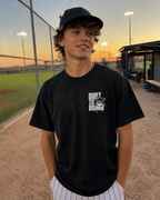 Teen at a baseball complex wearing a black Built to Bomb adult baseball T-shirt with the small white, hot pink and turquoise left-chest graphic.