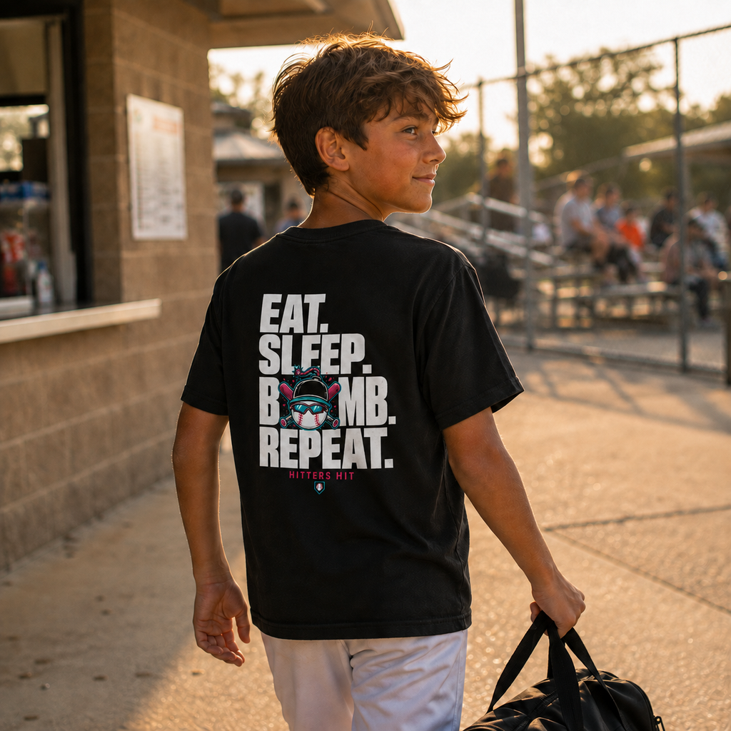 Preteen boy at a baseball field wearing a black youth baseball T-shirt with the large “Eat. Sleep. Bomb. Repeat.” back graphic in white, hot pink, and turquoise.