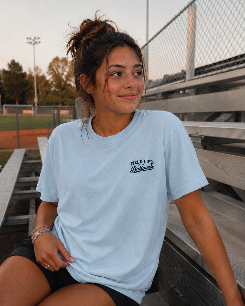 Teen girl wearing the chambray Late Nights at the Ballpark baseball T-shirt with a small front left chest graphic in a relaxed candid ballpark setting.