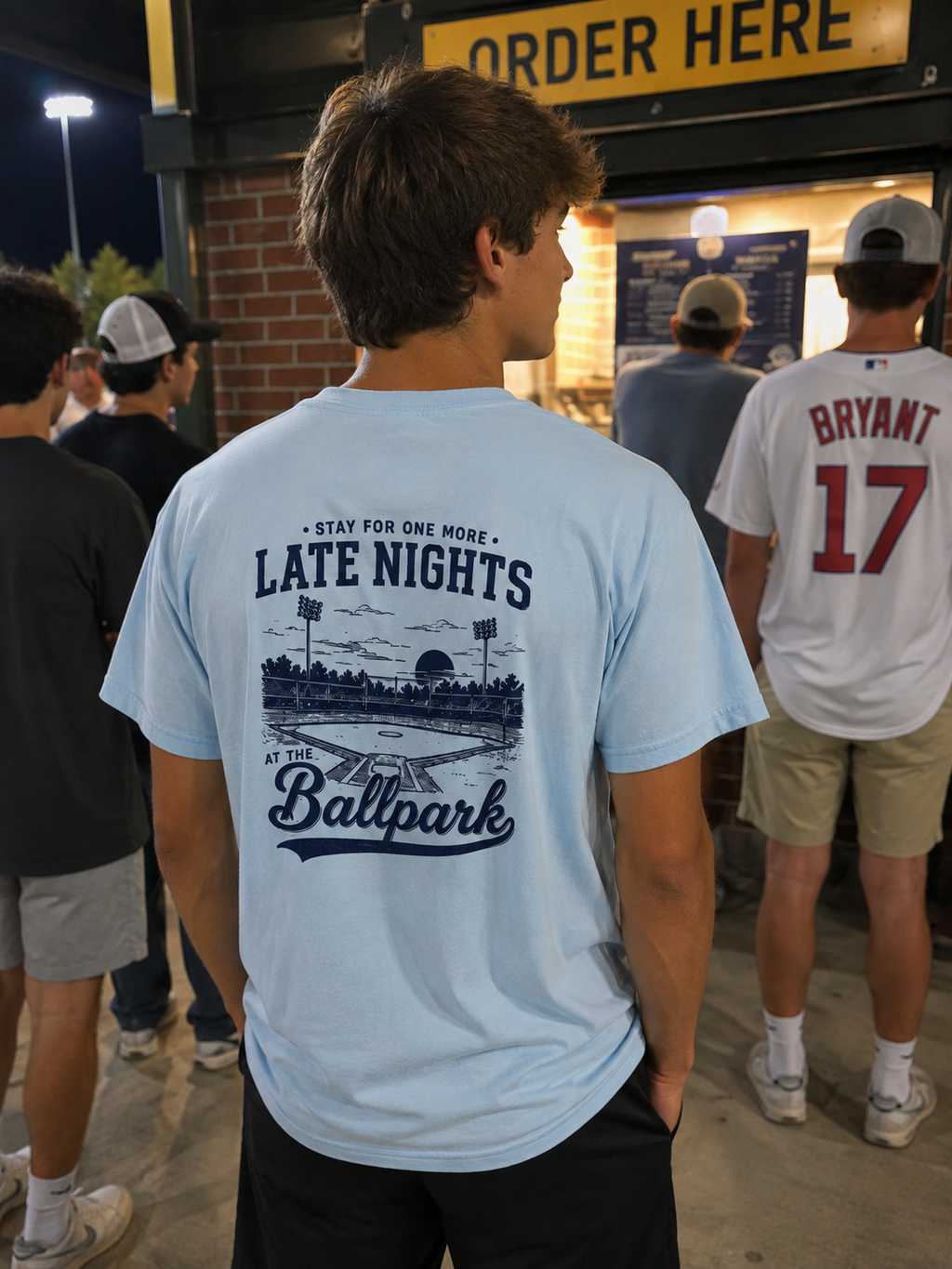 Teen wearing a Chambray Late Nights at the Ballpark baseball graphic T-shirt at a ballpark concession stand at night.