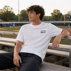 Teen wearing a white Late Nights at the Ballpark baseball T-shirt while sitting casually on bleachers at a baseball field.