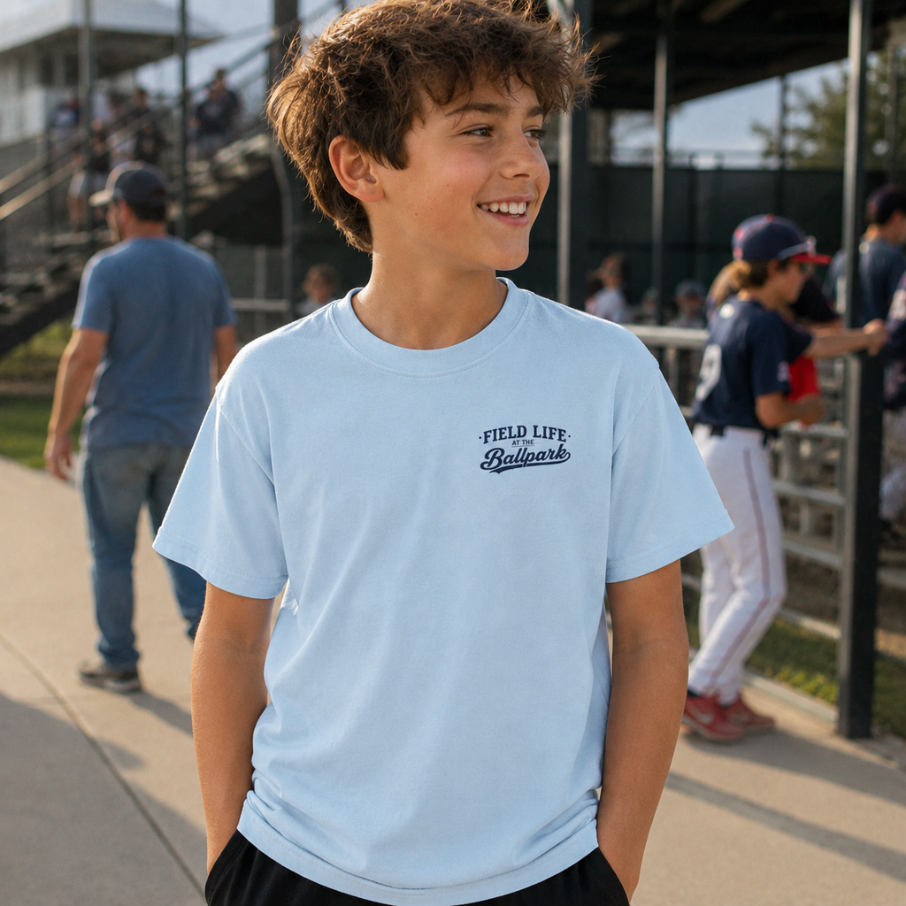 Boy wearing the chambray Late Nights at the Ballpark youth baseball T-shirt with small left chest design at a baseball field.