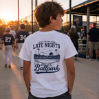 Boy wearing the white Late Nights at the Ballpark youth baseball T-shirt showing the large back graphic at a baseball field.