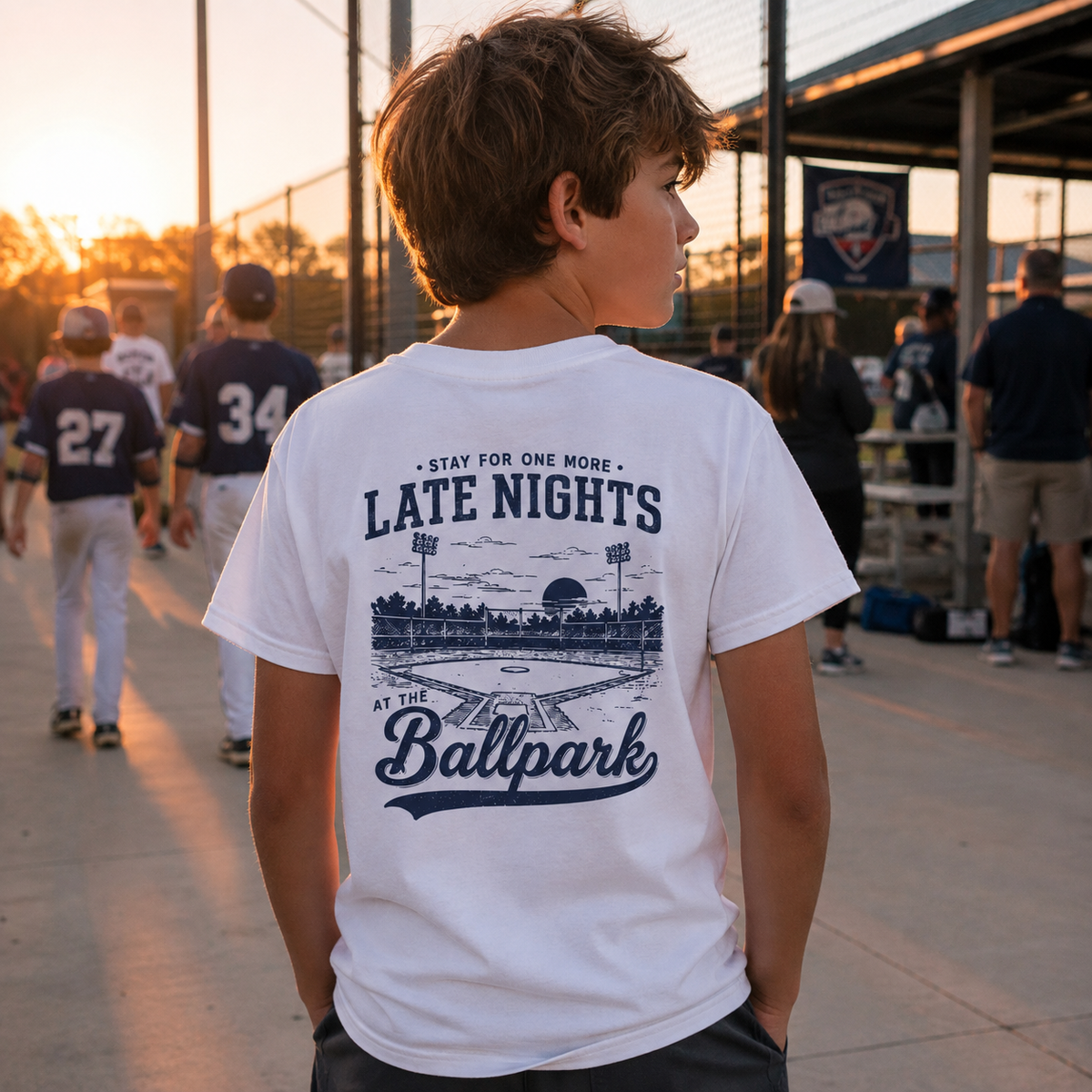 Boy wearing the white Late Nights at the Ballpark youth baseball T-shirt showing the large back graphic at a baseball field.