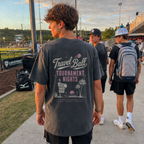 Teen boy wearing the Tournament Nights Palm Adult Baseball T-Shirt in Pepper, shown from the back at a baseball complex during golden hour with the large travel ball graphic visible.