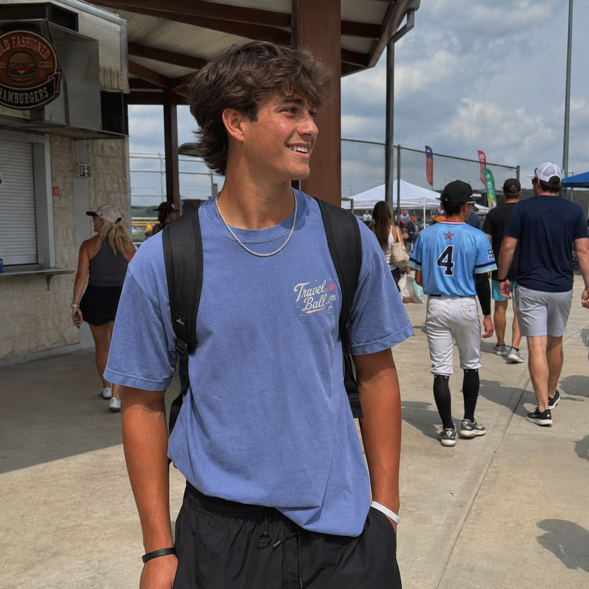 Teen boy wearing the Washed Denim adult baseball T-shirt at a baseball complex, showing the small Travel Ball left-chest graphic.