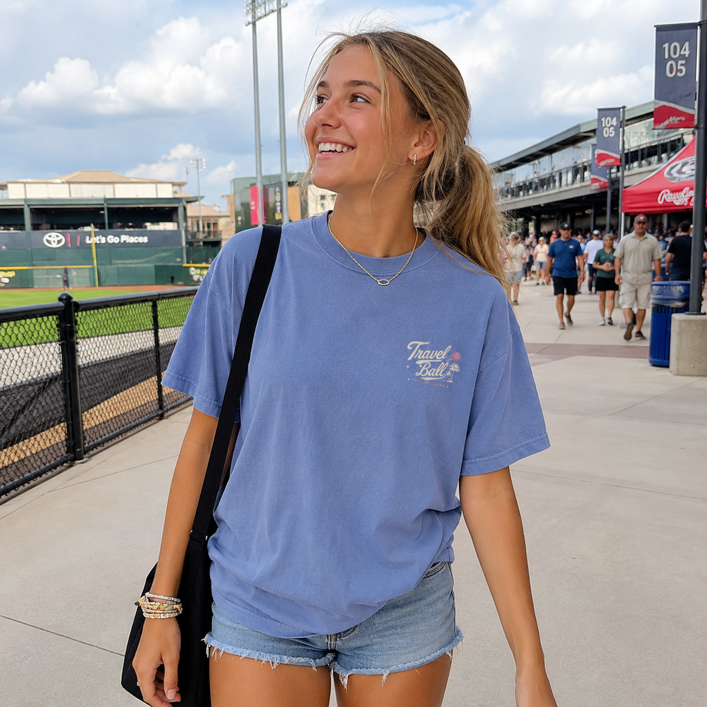 Teen girl wearing the Washed Denim adult baseball T-shirt at a ballpark, showing the small off-white and pink Travel Ball chest graphic.