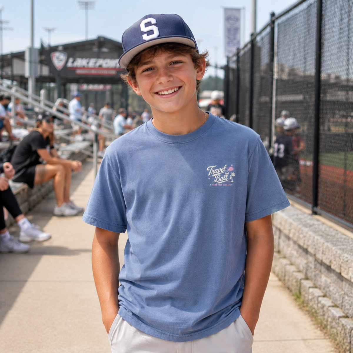 Preteen boy wearing the Washed Denim youth baseball T-shirt at a baseball complex, showing the small off-white and pink Travel Ball left-chest graphic.